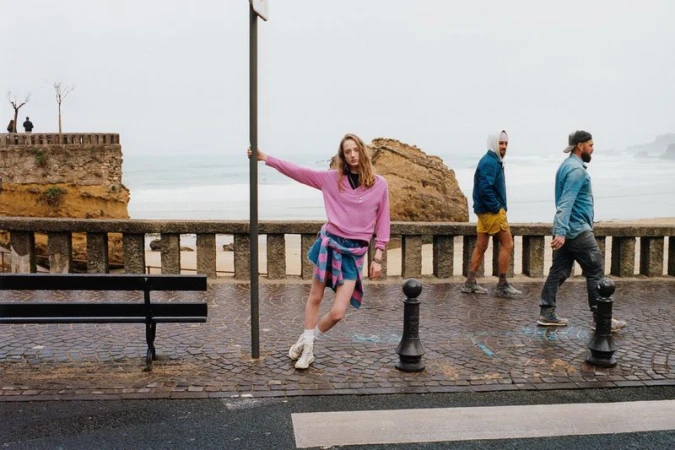 Woman poses by a pole near the ocean on a cloudy day, wearing a pink sweatshirt with two men walking behind her in the distance.