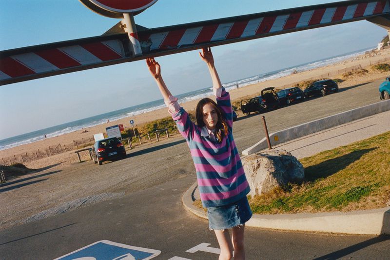Young woman in a striped shirt hanging from a striped bar at a beach parking lot with cars and the ocean in the background on a sunny day.