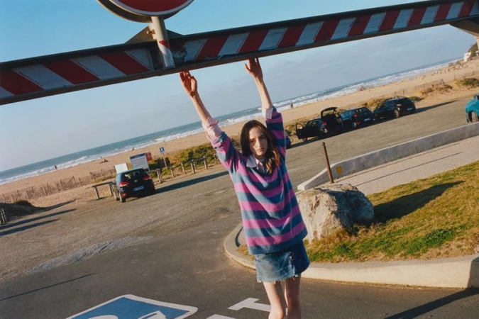 Young woman in a striped shirt hanging from a striped bar at a beach parking lot with cars and the ocean in the background on a sunny day.