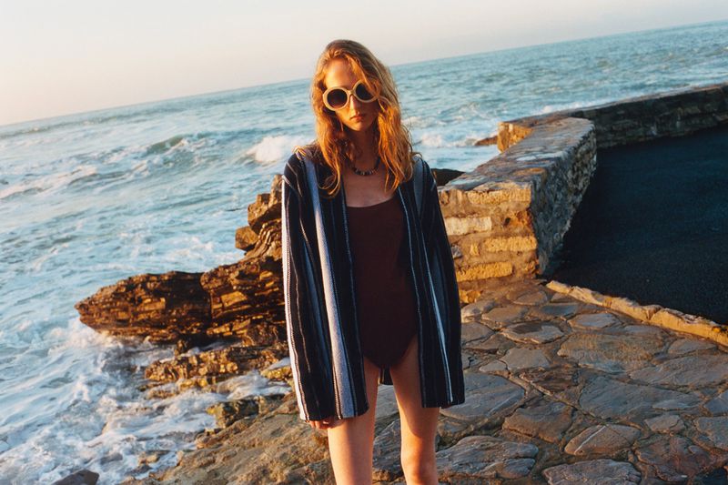 A woman stands on a stone platform near the ocean, wearing sunglasses and a striped cover-up over a dark swimsuit at sunset.