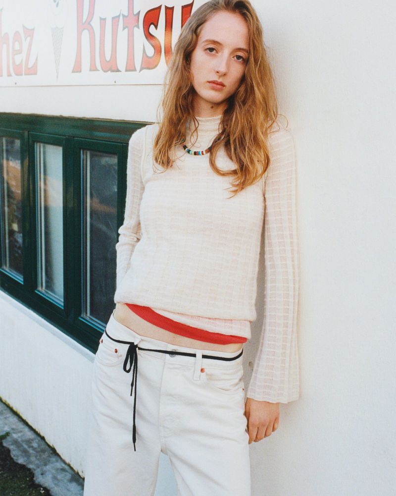 A fair-skinned woman with long red hair poses against a white wall with a window, wearing a cream top and white jeans with a black string belt.