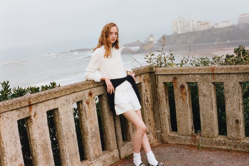 A young adult female model poses on a concrete ledge by the ocean, wearing a white blouse and shorts, with buildings visible in the background.
