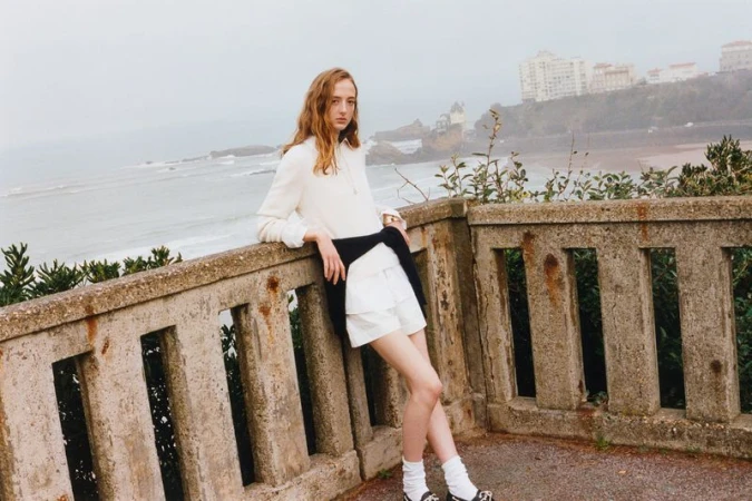 A young adult female model poses on a concrete ledge by the ocean, wearing a white blouse and shorts, with buildings visible in the background.