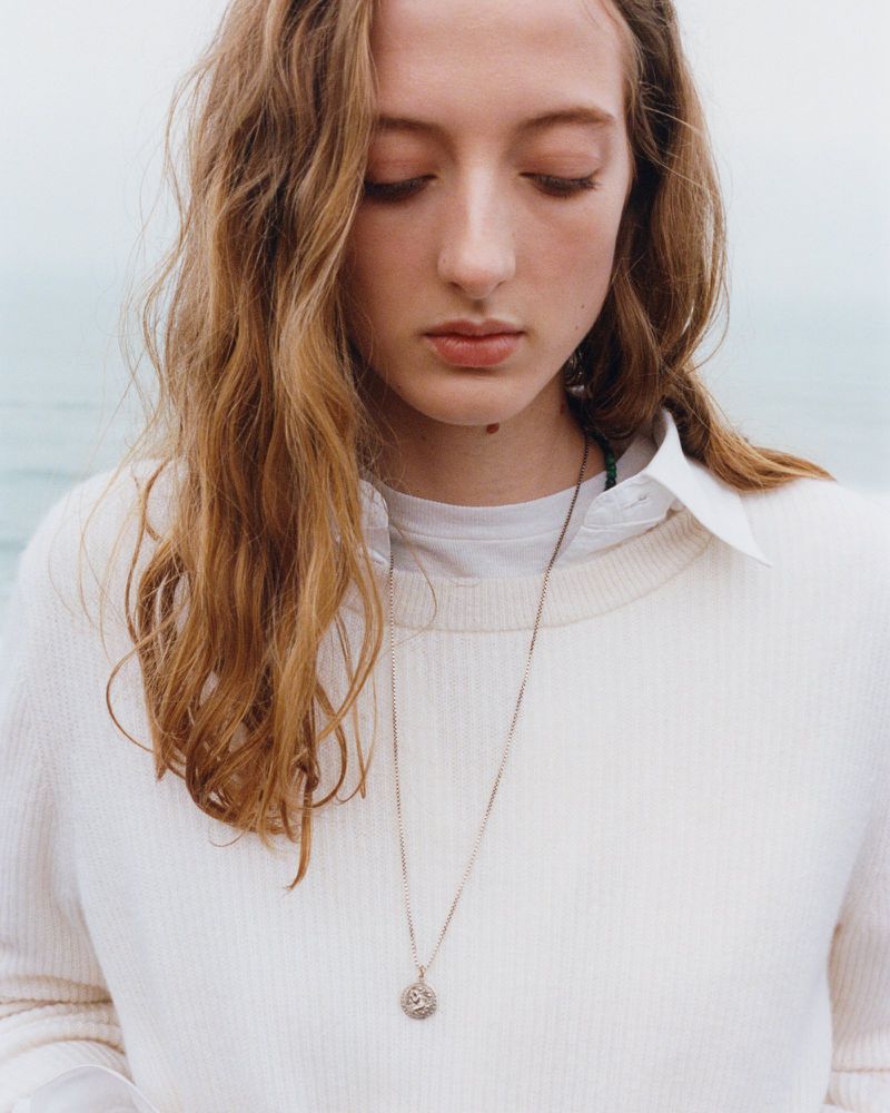 Young woman with wavy hair, wearing a white sweater and a delicate pendant necklace, poses in a natural outdoor setting.