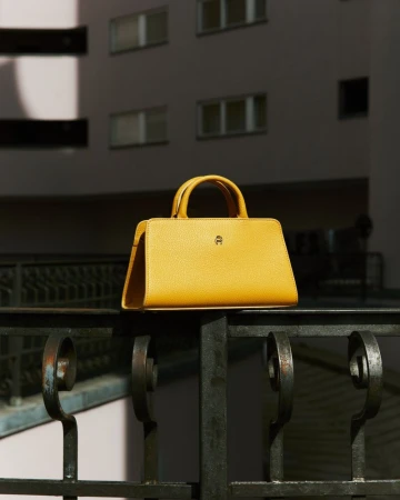 A yellow Aigner handbag sits on a wrought-iron railing, photographed in front of a light pink building with windows, outdoors.