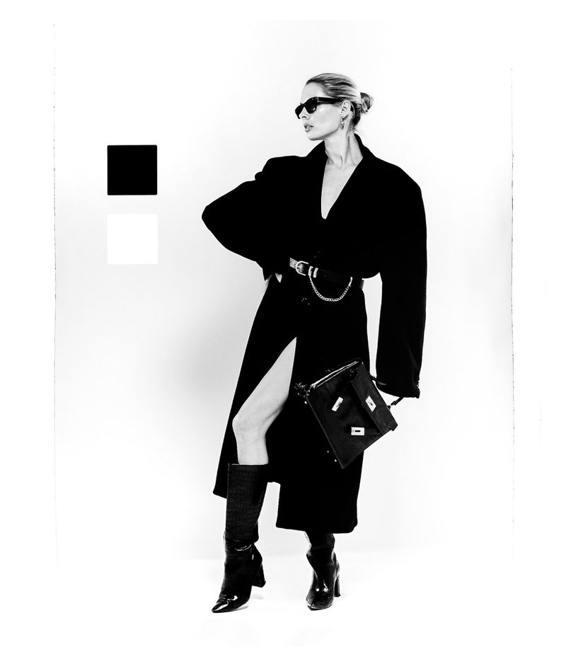 Black and white fashion shot of a female model in a long black coat, sunglasses, boots, and a handbag against a white background.