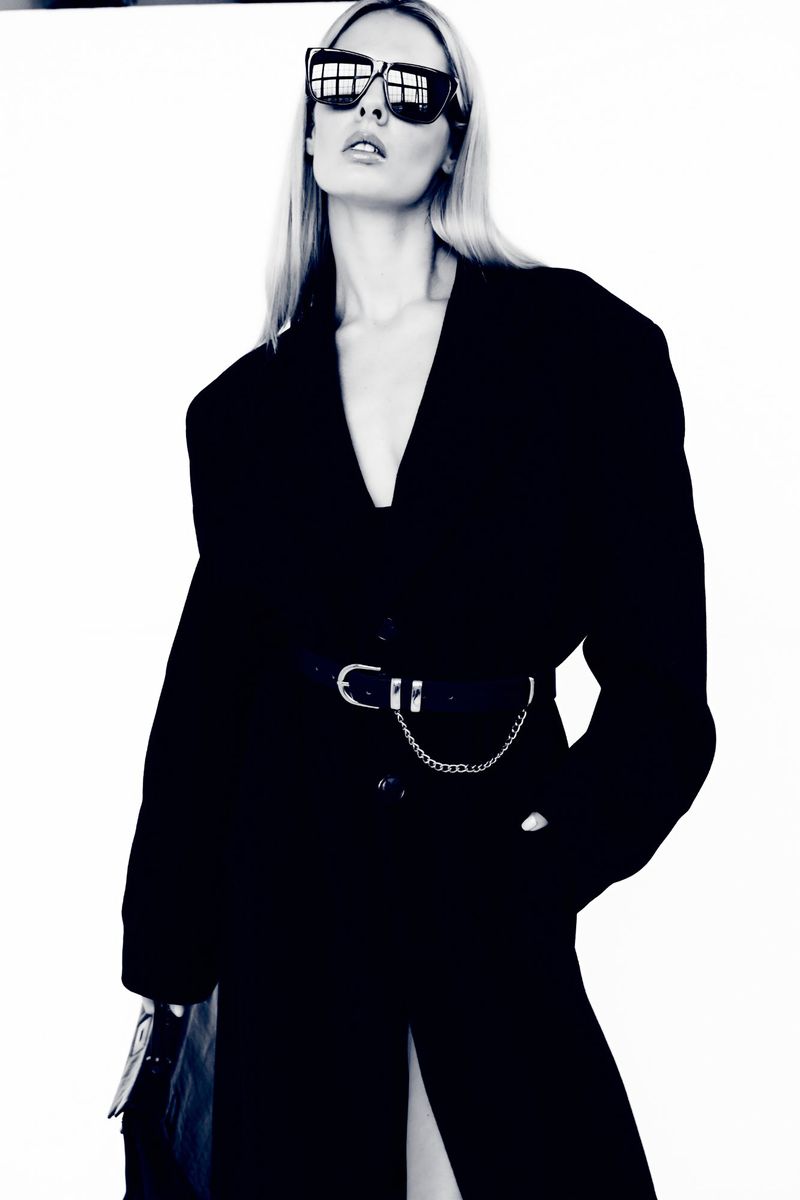 A blonde female model poses indoors against a white backdrop, wearing a black coat, dark sunglasses, and a stylish chain belt.