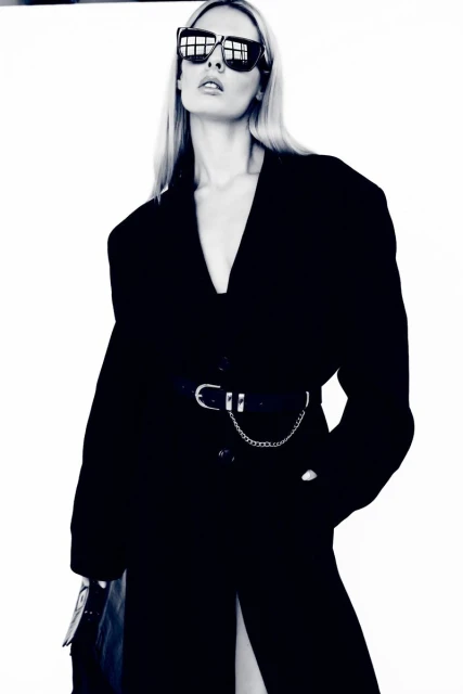 A blonde female model poses indoors against a white backdrop, wearing a black coat, dark sunglasses, and a stylish chain belt.