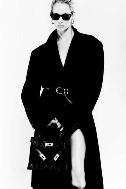 Black and white portrait of a fashionable woman in sunglasses and an overcoat, holding a leather purse against a white background.