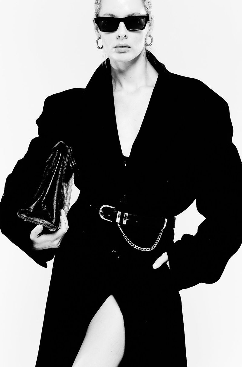 A striking black-and-white portrait of a fashion model in a black jacket, dark sunglasses, and hoop earrings, holding a clutch. 