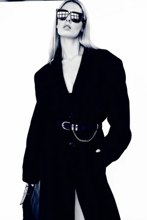 Model Emily Fay wearing dark sunglasses and a black belted coat against a plain white studio background.