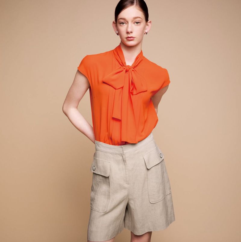 Elegant young woman in orange bow blouse and beige cargo shorts poses in a fashion studio with soft, neutral background lighting.