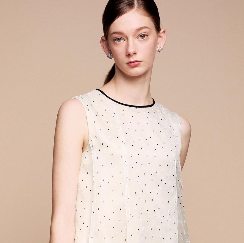 Portrait of a poised young woman with elegant earrings, wearing a sleeveless polka dot top against a light beige background.