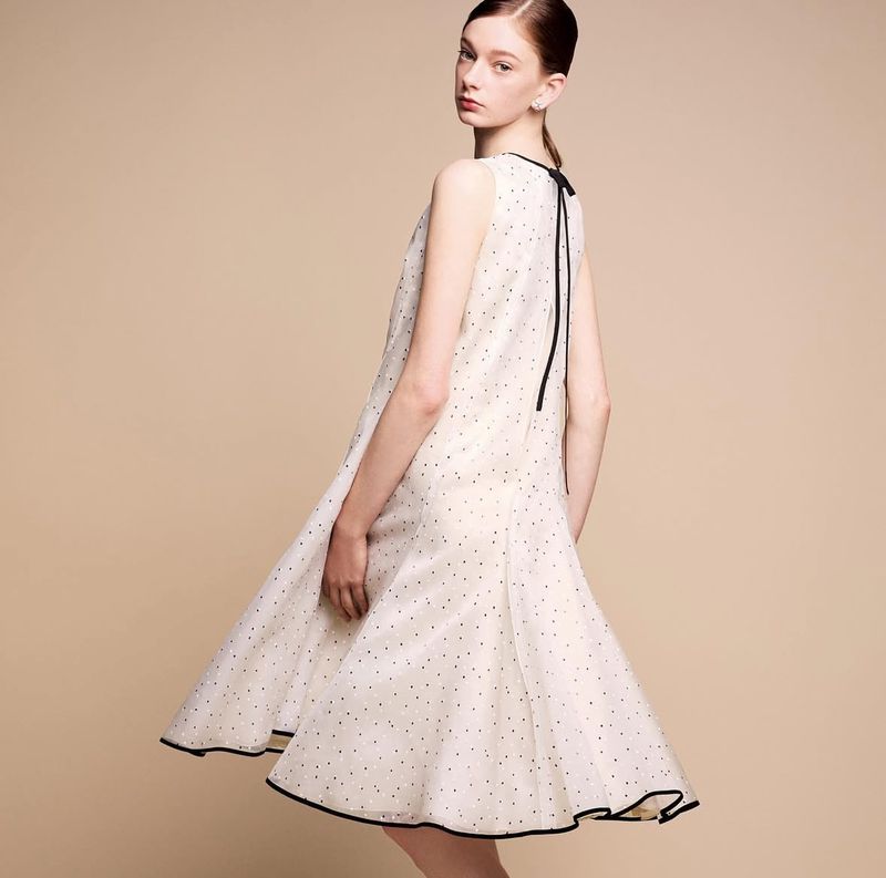 Fashion shot of a young woman wearing a white dress with black polka dots, posing in a studio setting with a beige background.