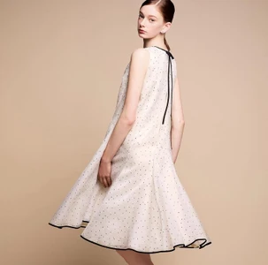 Model in white polka dot sleeveless dress with black trim standing against a seamless beige studio background.