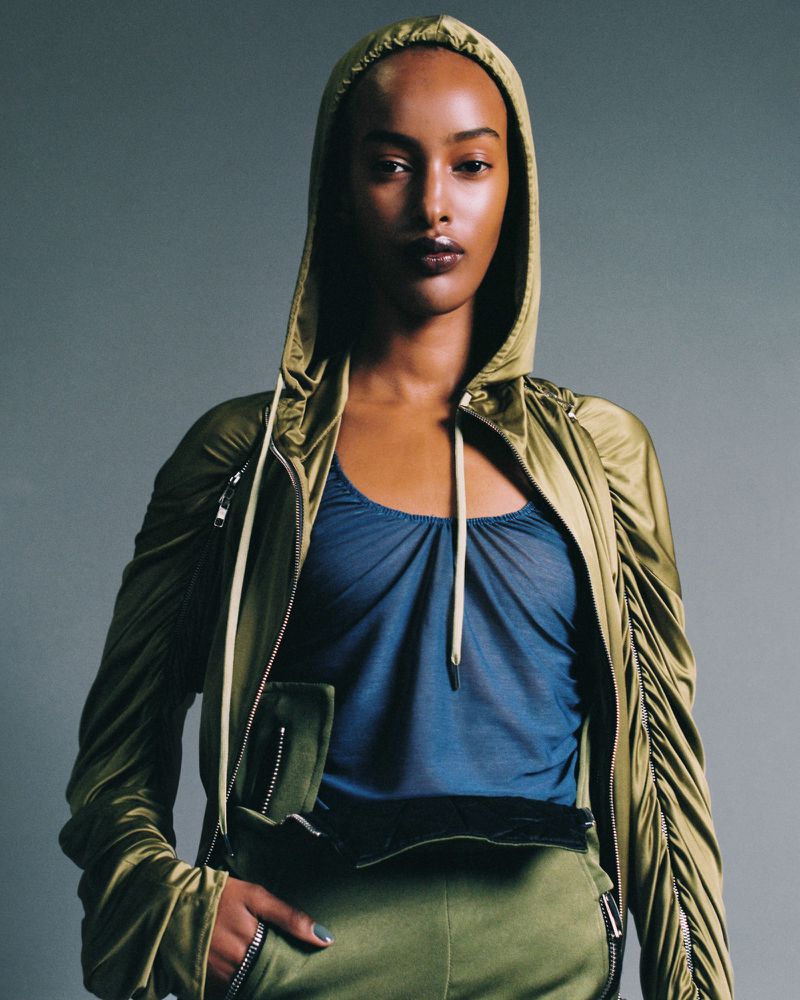 Portrait of a slender Black woman in a hooded olive jacket and blue top, hand on hip in olive pants, against a grey backdrop.