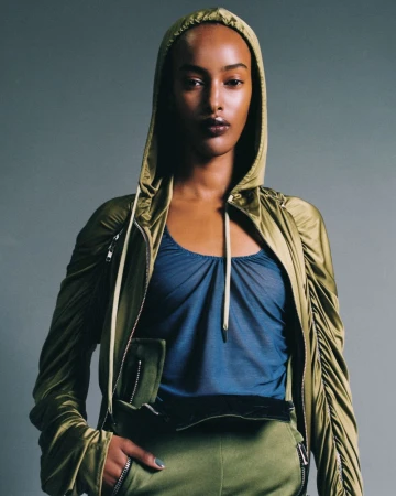 Portrait of a slender Black woman in a hooded olive jacket and blue top, hand on hip in olive pants, against a grey backdrop.