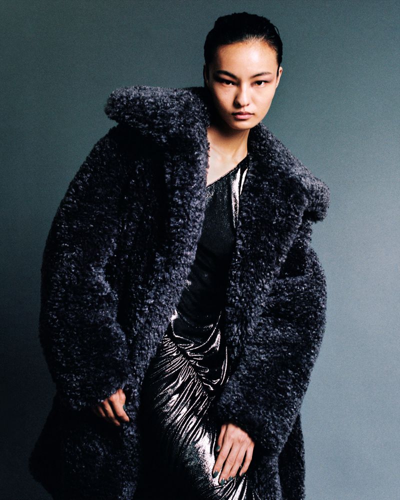 Fashion shot of a woman in a long, oversized, fluffy dark grey coat and a sparkly silver dress, posing against a grey backdrop.