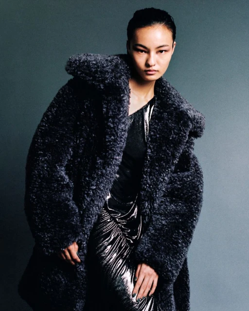 Fashion shot of a woman in a long, oversized, fluffy dark grey coat and a sparkly silver dress, posing against a grey backdrop.