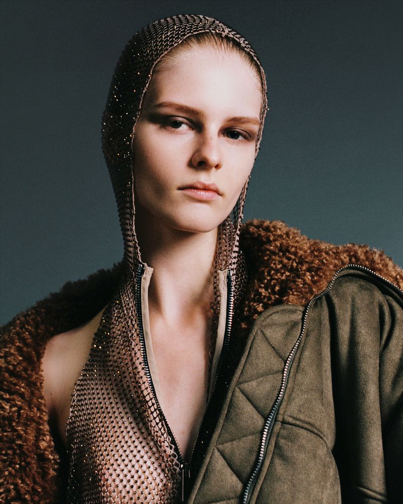 A woman in a metallic hooded top and brown fur coat poses against a gray backdrop in a fashion-style studio portrait.