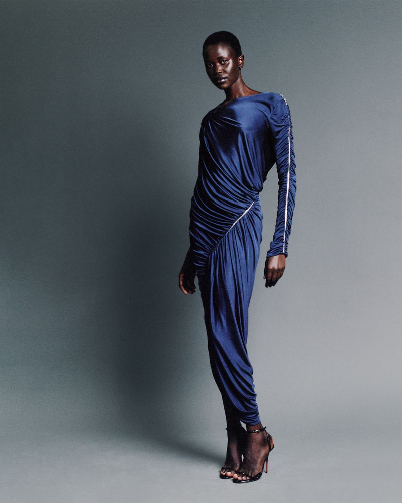 Model wearing a blue draped long-sleeved dress and black heeled sandals posing against a grey seamless studio background.