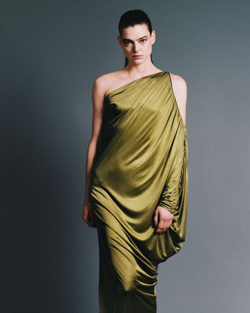 Elegant woman poses in a flowing, olive green, one-shoulder dress against a neutral gray backdrop in a fashion photography studio.