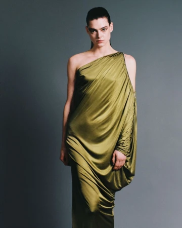 Elegant woman poses in a flowing, olive green, one-shoulder dress against a neutral gray backdrop in a fashion photography studio.