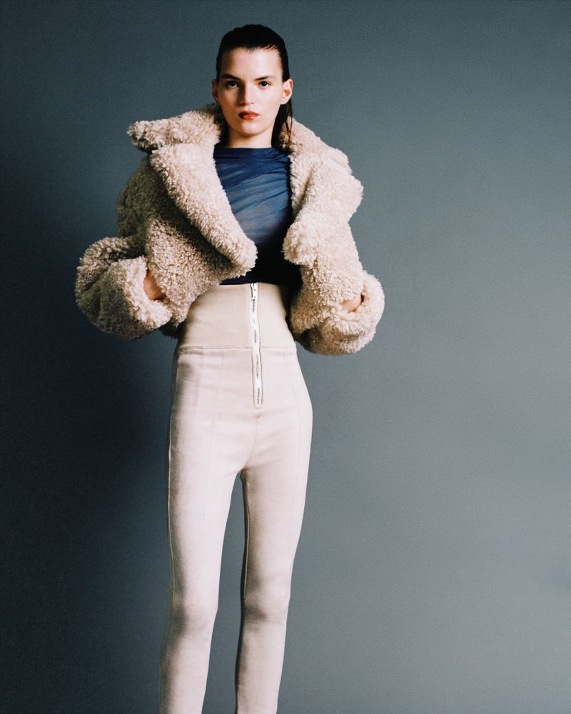 Model wearing a fuzzy cream jacket and high-waisted trousers against a solid grey studio background.