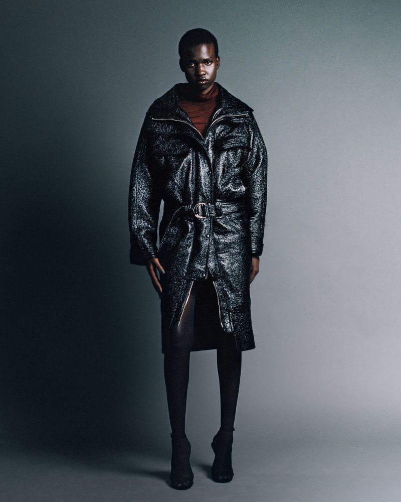 Model wearing a black glossy belted coat and dark stockings standing against a gradient studio background.