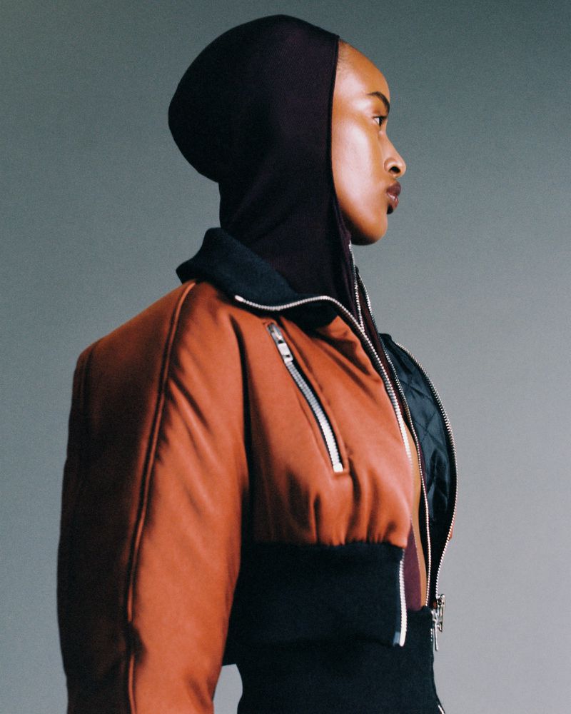 Profile portrait of an African American woman in a rust-colored bomber jacket with a black hood against a gray background in studio lighting.