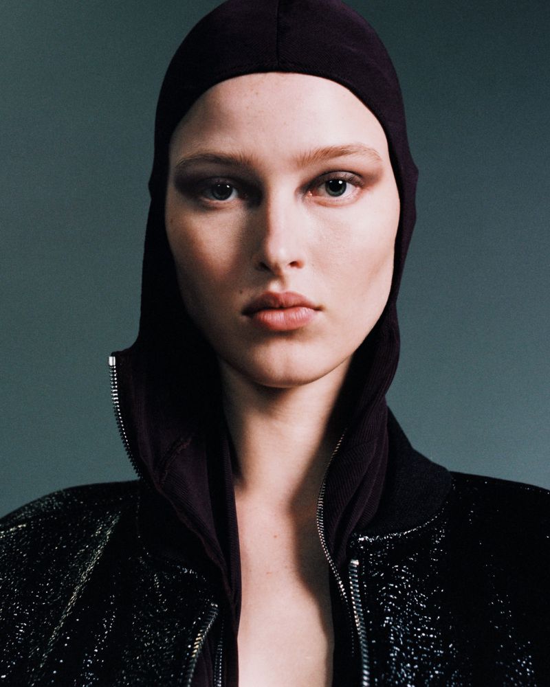 Close-up portrait of a model wearing a dark hooded top and a glittering jacket against a neutral studio background