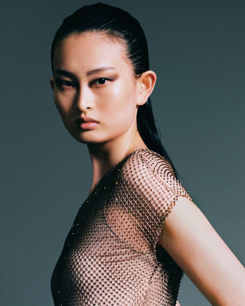 Portrait of a woman with her hair pulled back wearing a textured metallic top against a grey background in a studio setting.
