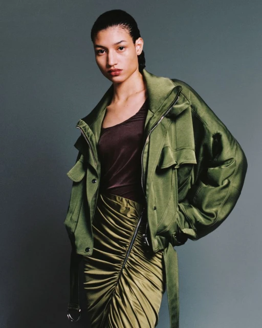 Fashion portrait of a woman in a green jacket and ruched skirt, posing in a studio with a neutral background. The image has studio lighting.
