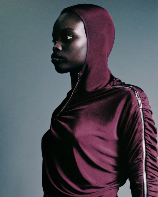 Portrait of a dark-skinned woman in profile wearing a hooded, long-sleeved purple dress with silver zippers on the side. Studio shot.