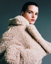 Model wearing a textured cream shearling coat against a dark grey seamless studio background.