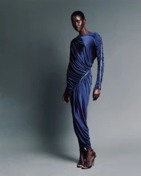 Model wearing a blue draped long-sleeved dress and black heeled sandals posing against a grey seamless studio background.