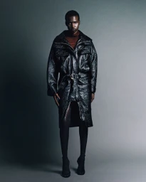 Model wearing a black glossy belted coat and dark stockings standing against a gradient studio background.