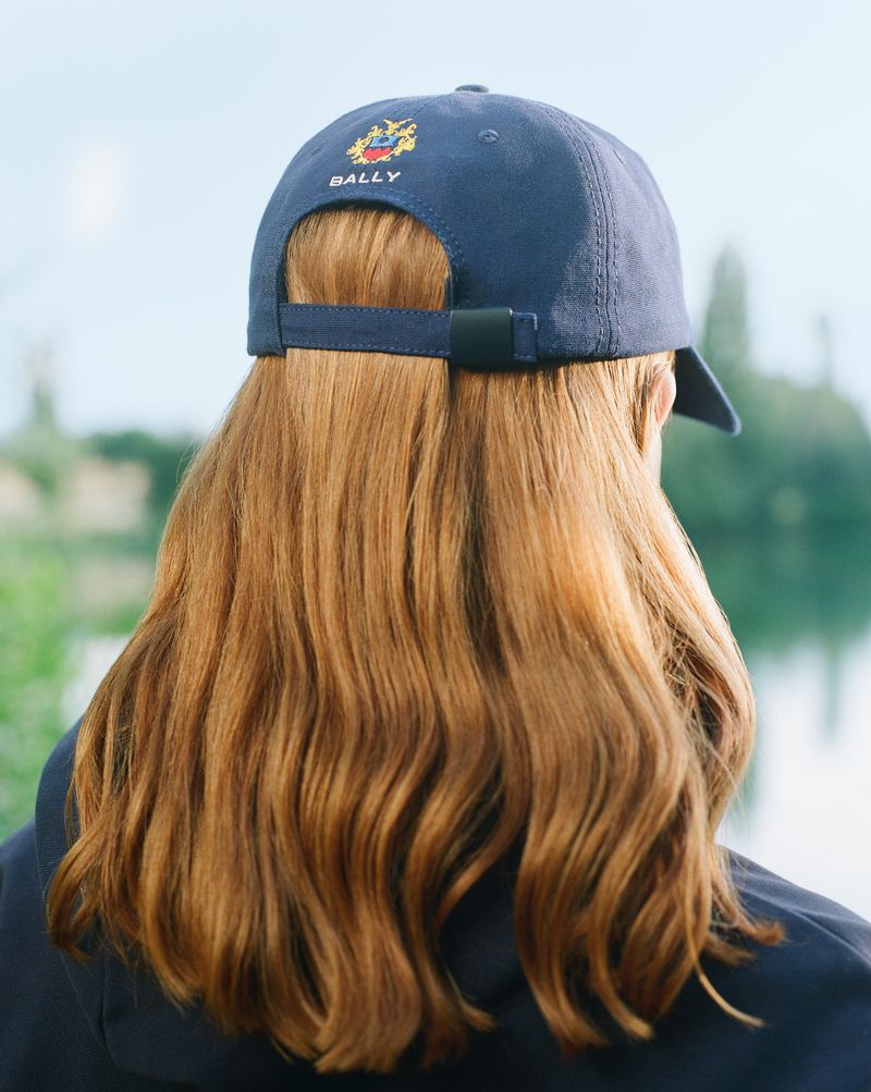Person wearing a navy Bally baseball cap with auburn wavy hair, viewed from behind in an outdoor setting with trees and water.