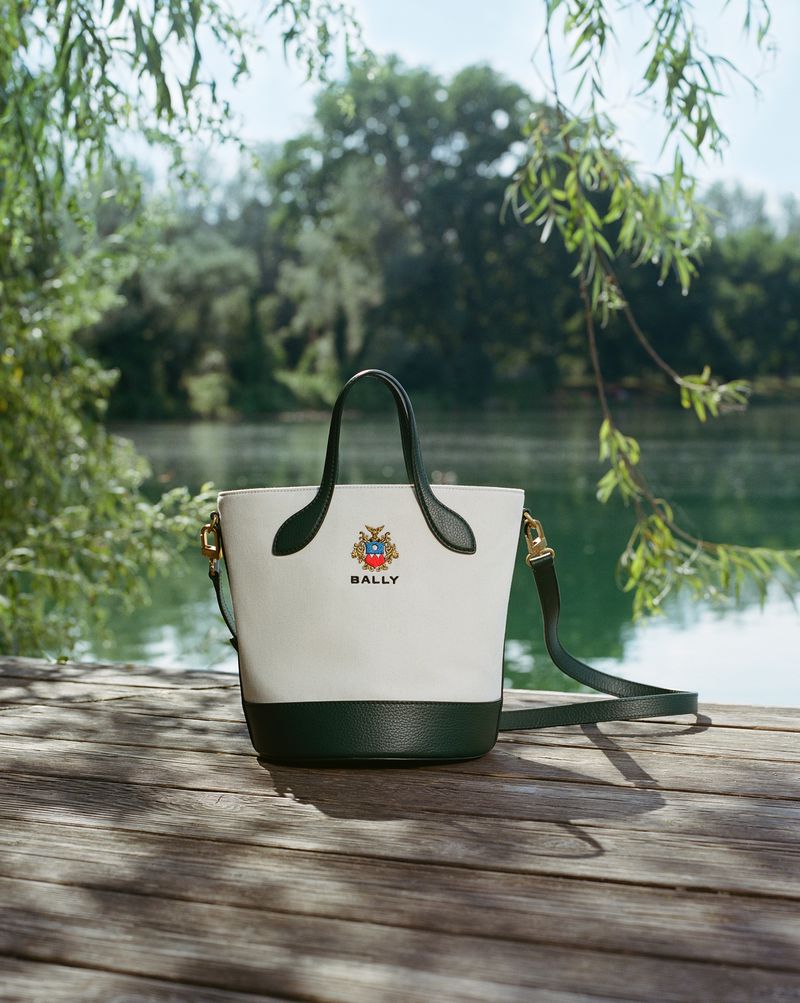 Bally bucket bag in white and dark green leather sits on wooden planks in front of a lake with green trees and foliage.