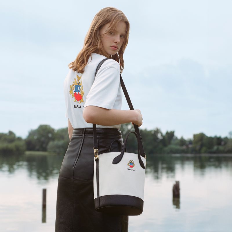 Woman in white tee and leather skirt carrying a two-tone Bally bucket bag outdoors near a reflective body of water and greenery.