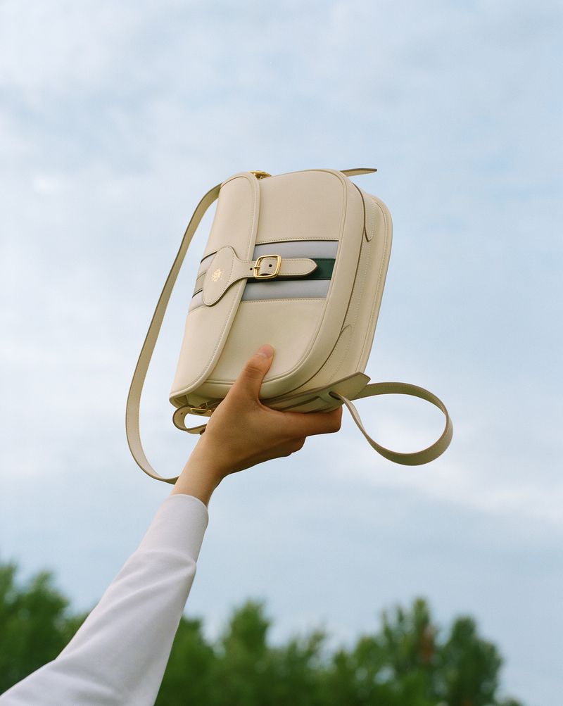 Cream-colored leather satchel with a striped band held up by a hand wearing a white shirt against a sky and treeline backdrop.