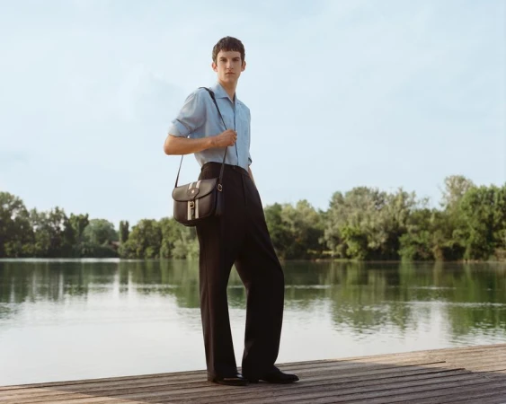 A young man stands on a wooden dock by a lake with trees in the background, wearing a blue shirt, black trousers, and a shoulder bag.