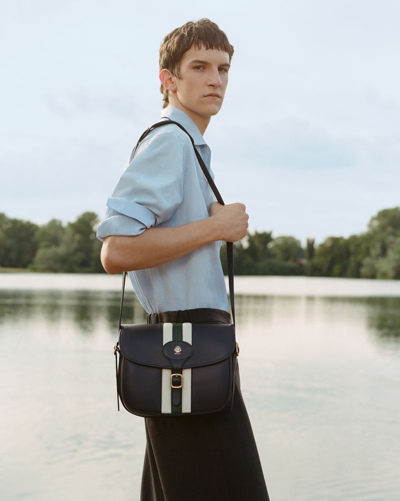 A young adult male stands near a lake wearing a blue button-down shirt and a black crossbody bag with green and white stripes.
