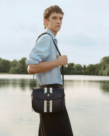 A young adult male stands near a lake wearing a blue button-down shirt and a black crossbody bag with green and white stripes.