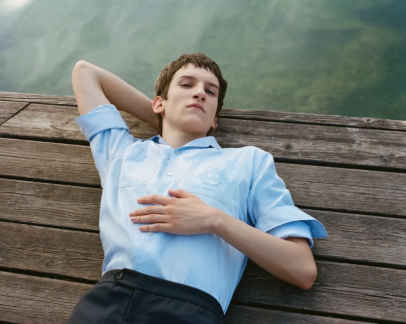 A young adult lies on a wooden dock with a blue shirt and dark pants in the sunlight, with water visible in the background.