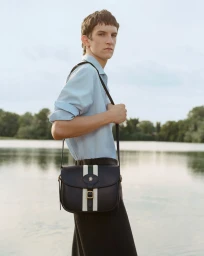 Man wearing a light blue shirt carrying a dark leather crossbody bag with white vertical stripes by a calm lake.