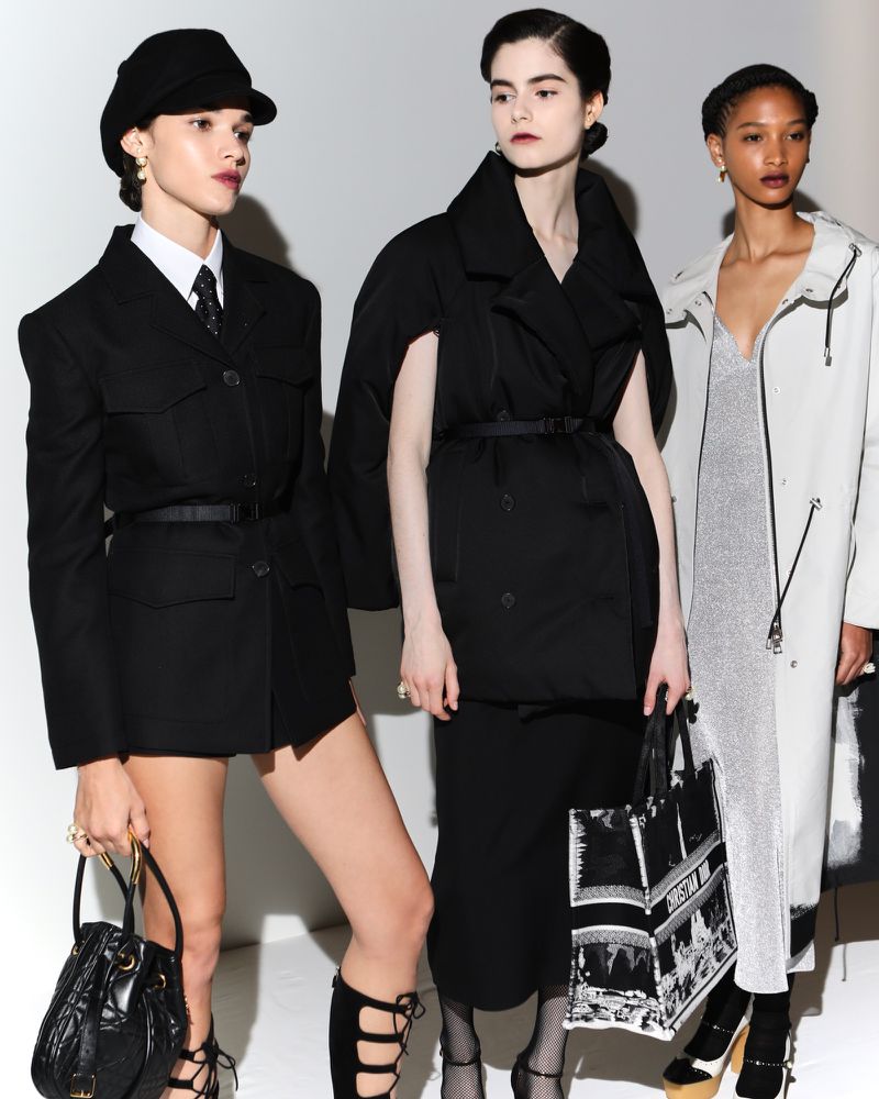 Three fashion models pose in a studio against a white backdrop. The models wear black and white outfits and hold handbags.