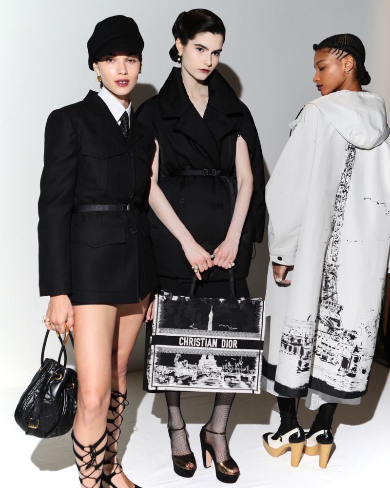 Three fashion models pose in black and white outfits against a white backdrop, one holding a Christian Dior tote bag.
