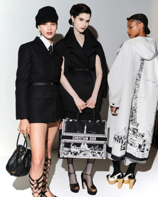 Three fashion models pose in black and white outfits against a white backdrop, one holding a Christian Dior tote bag.