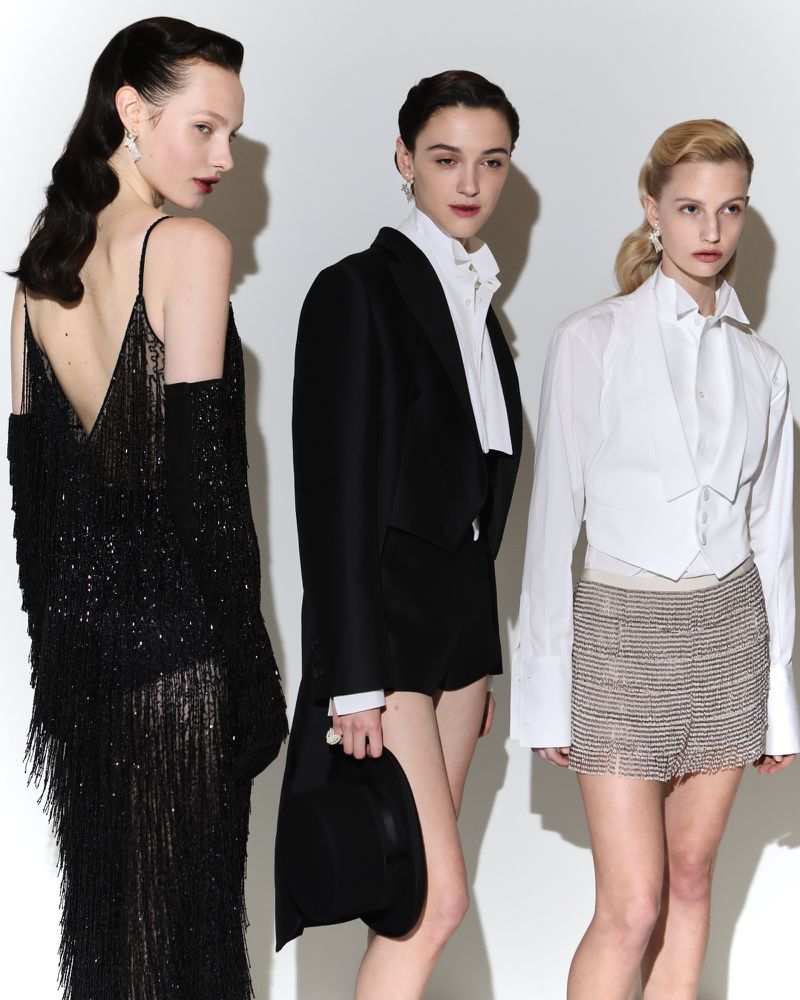 Three slender fashion models in chic evening wear, including a fringe dress and tailored suits, pose against a stark white backdrop.